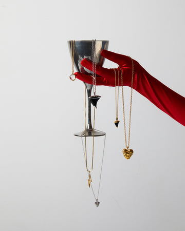 Hand in a red glove holding a martini glass with hanging gold necklaces on a light gray background