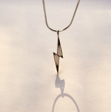 A smokey quartz lightning bolt pendant necklace hanging from a thin silver snake chain against a plain off-white background, casting a sharp shadow below.