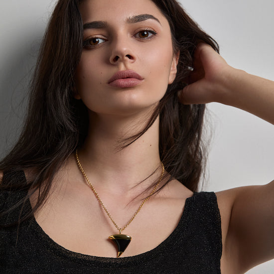 Woman wearing a  gold necklace with an onxy stone. shaped like a thorn against a neutral background