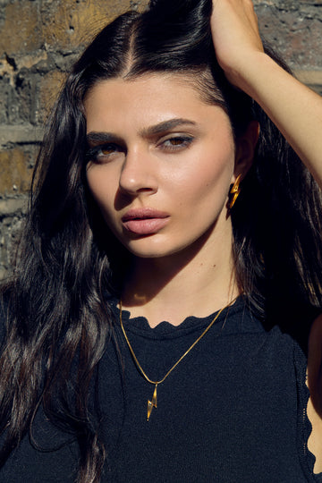 Woman with long dark hair and gold jewelry against a brick wall.
