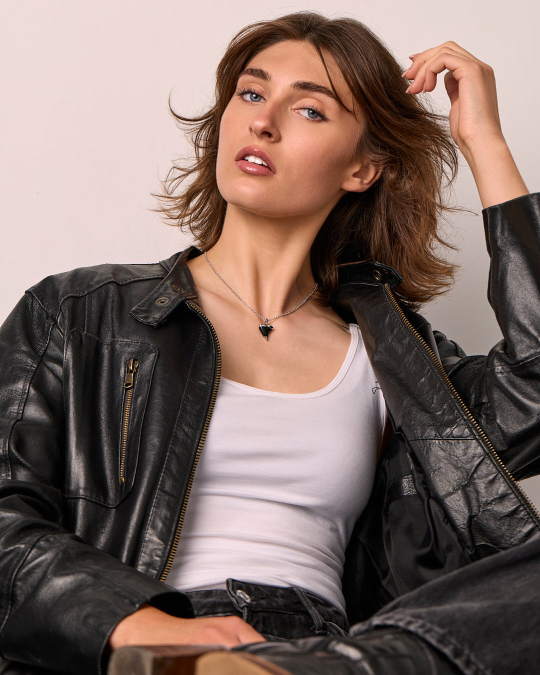 Woman wearing a black leather jacket over a white top, sitting against a plain background