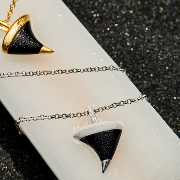 A close-up of a necklace with a conical pendant featuring a black glitter center, a white outer section, and a silver-toned pointed tip, shown on a white display bar against a dark glittery background.
