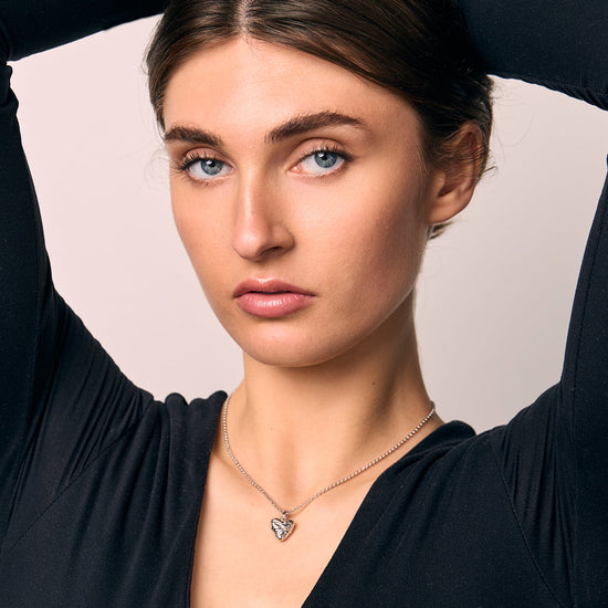 Woman wearing a black top and small heart pendant with her arms raised, against a plain background