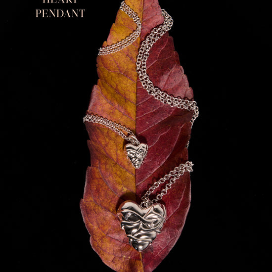Silver small heart pendant and large heart locket both with thorns wrapped around the pendant resting on an autumnal leaf and a black background.